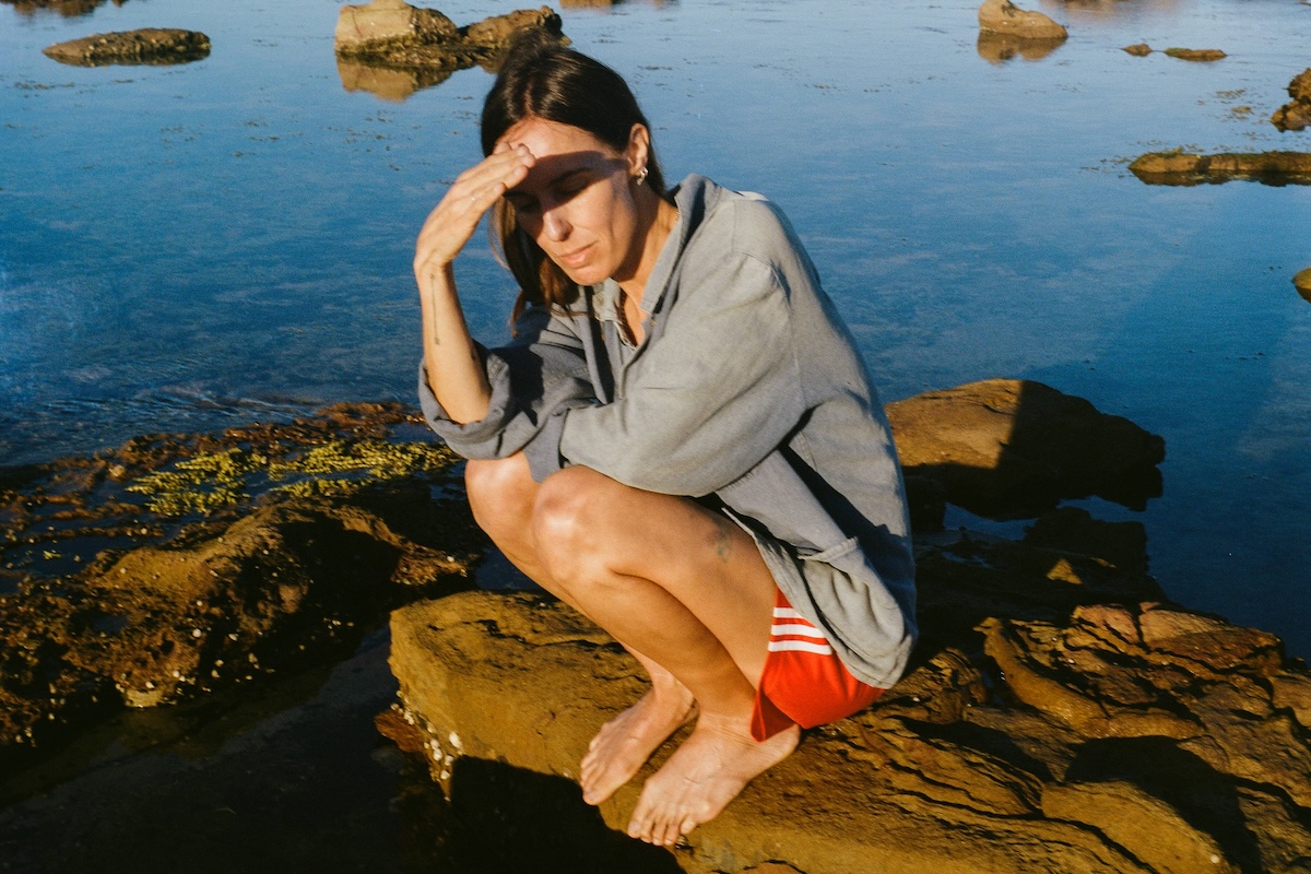Musician Holly Throsby crouching barefoot on coastal rocks, shielding her eyes from the sun wearing red shorts and a denim jacket.